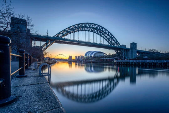 The Tyne Bridge reflected in the River Tyne beside the quayside at dusk.